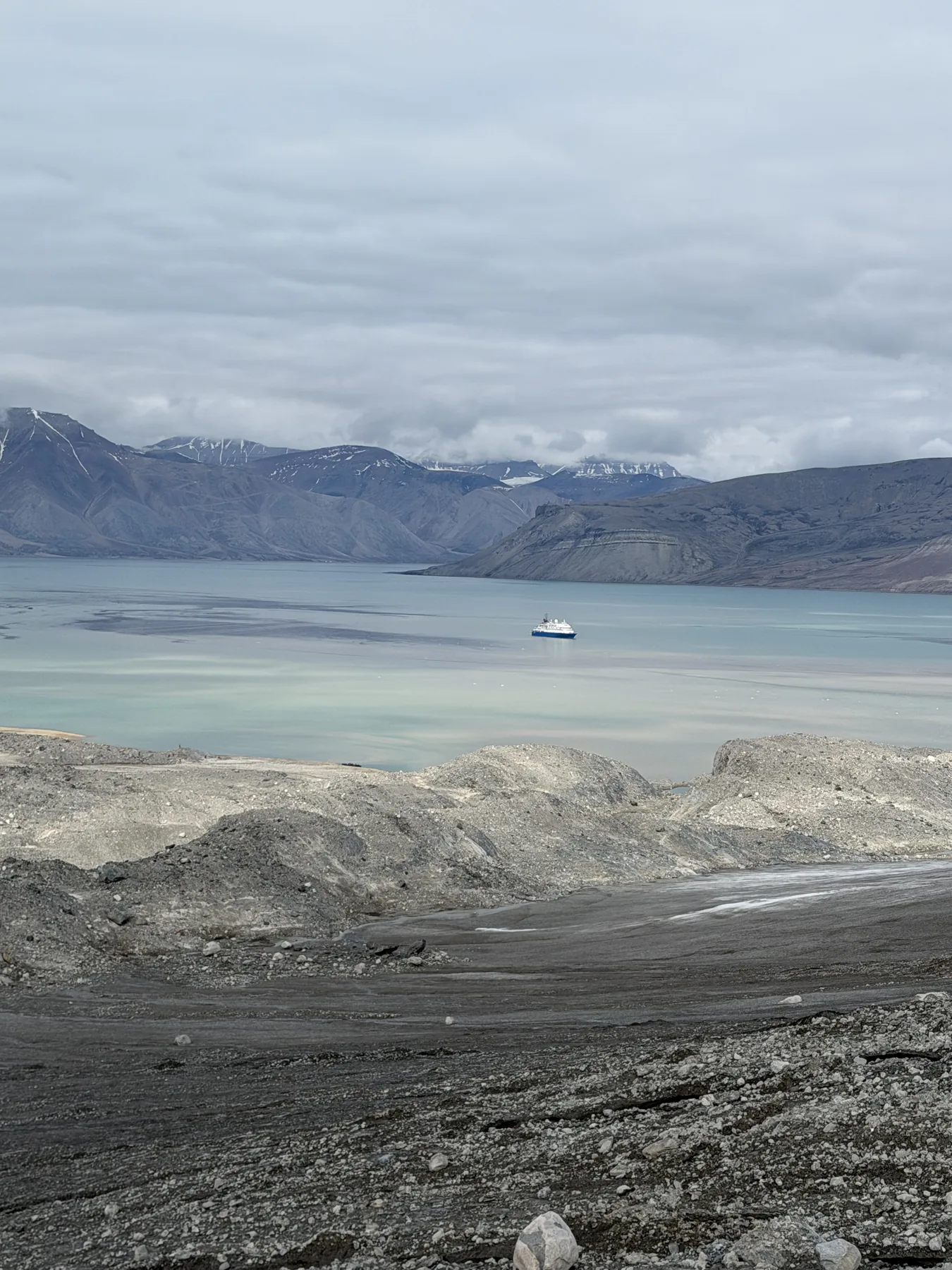 Moraine rubble in foreground, glacial-milk fjord beyond, a ship at anchor for scale